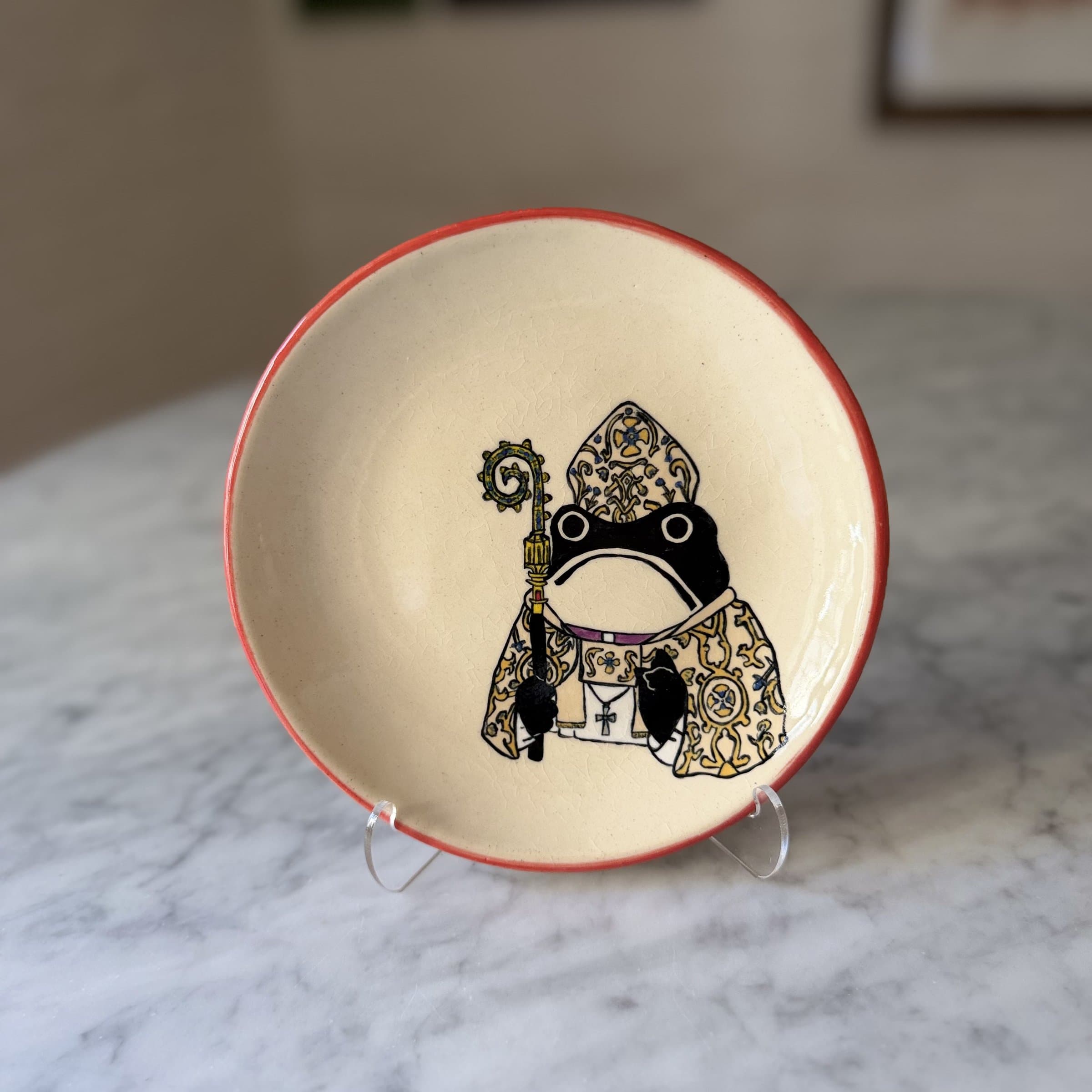 A hand-painted plate of a frog in pope vestments and mitre.