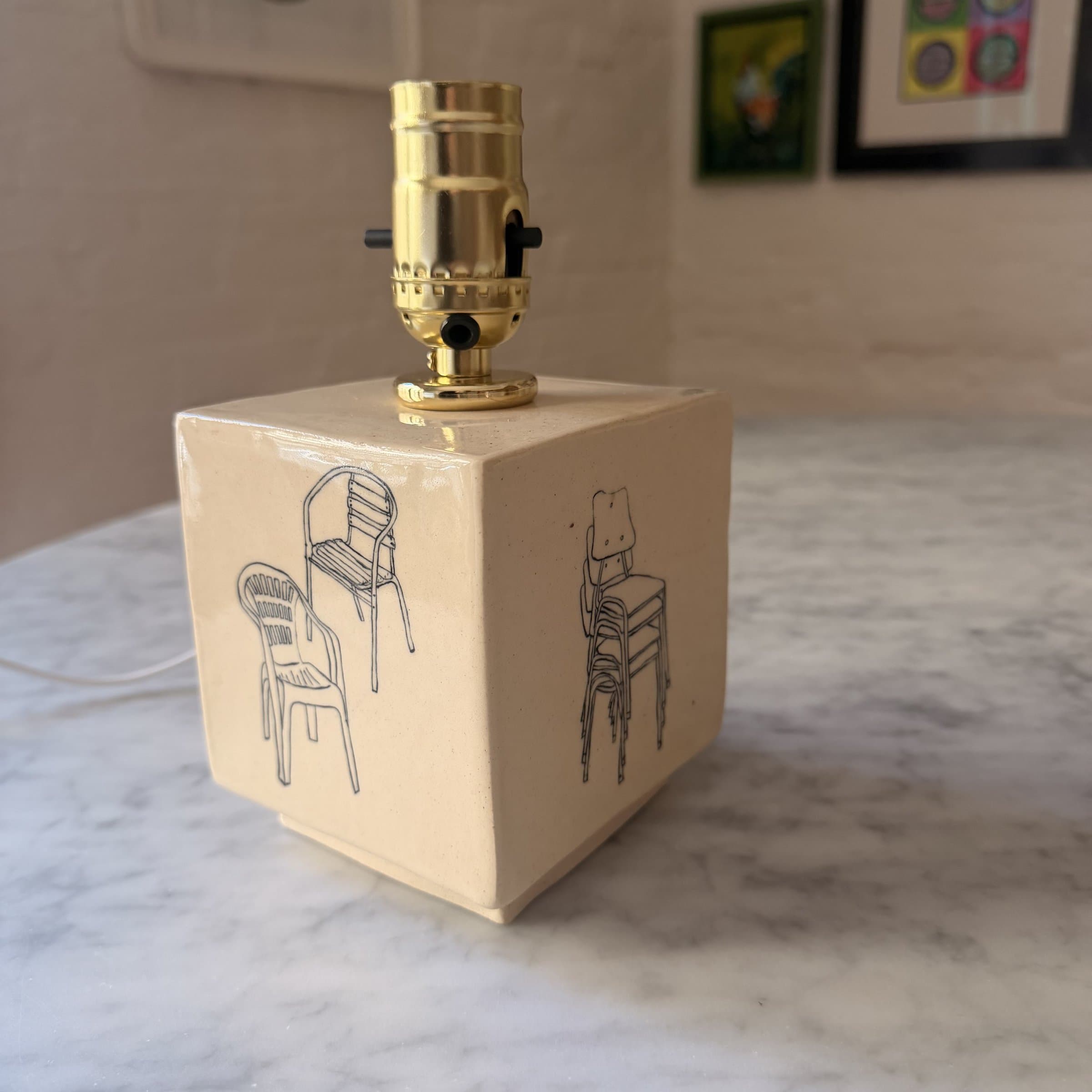 A cube-shaped ceramic lamp base drawn with plastic patio chairs on each side.
