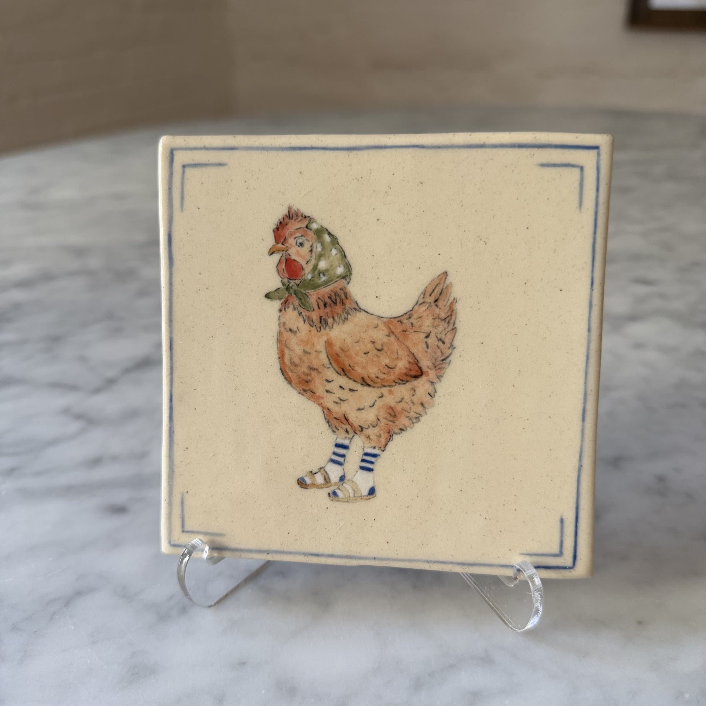A hand-painted tile of a hen wearing blue striped socks and sandals, with a polka-dot head scarf.