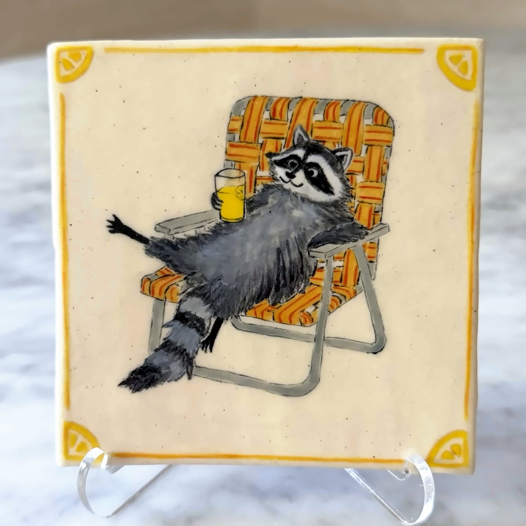 A tile painted with a raccoon lounging in a woven lawn chair, holding a glass of lemonade.