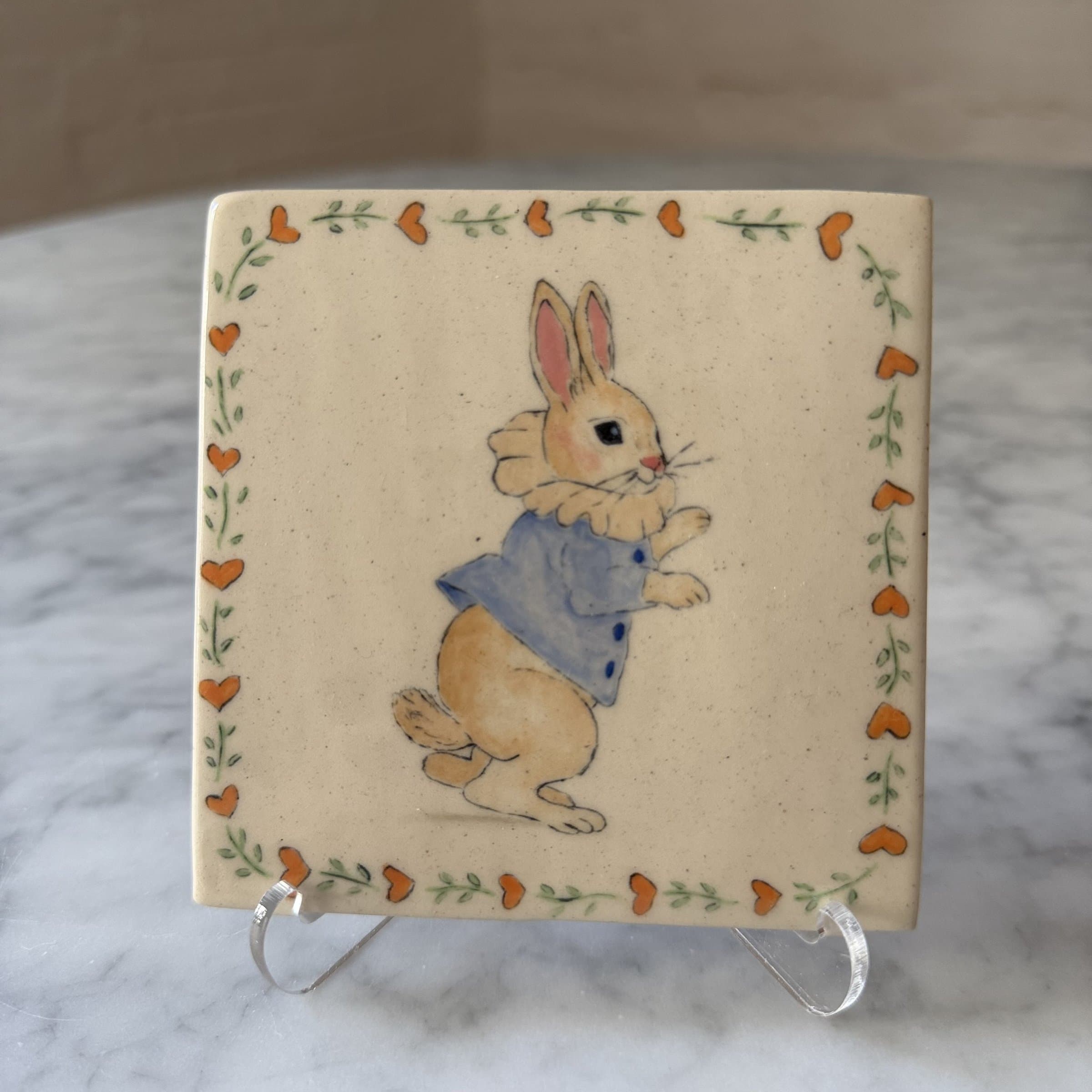 A hand-painted tile of a rabbit in the Beatrix Potter style, in a blue coat.
