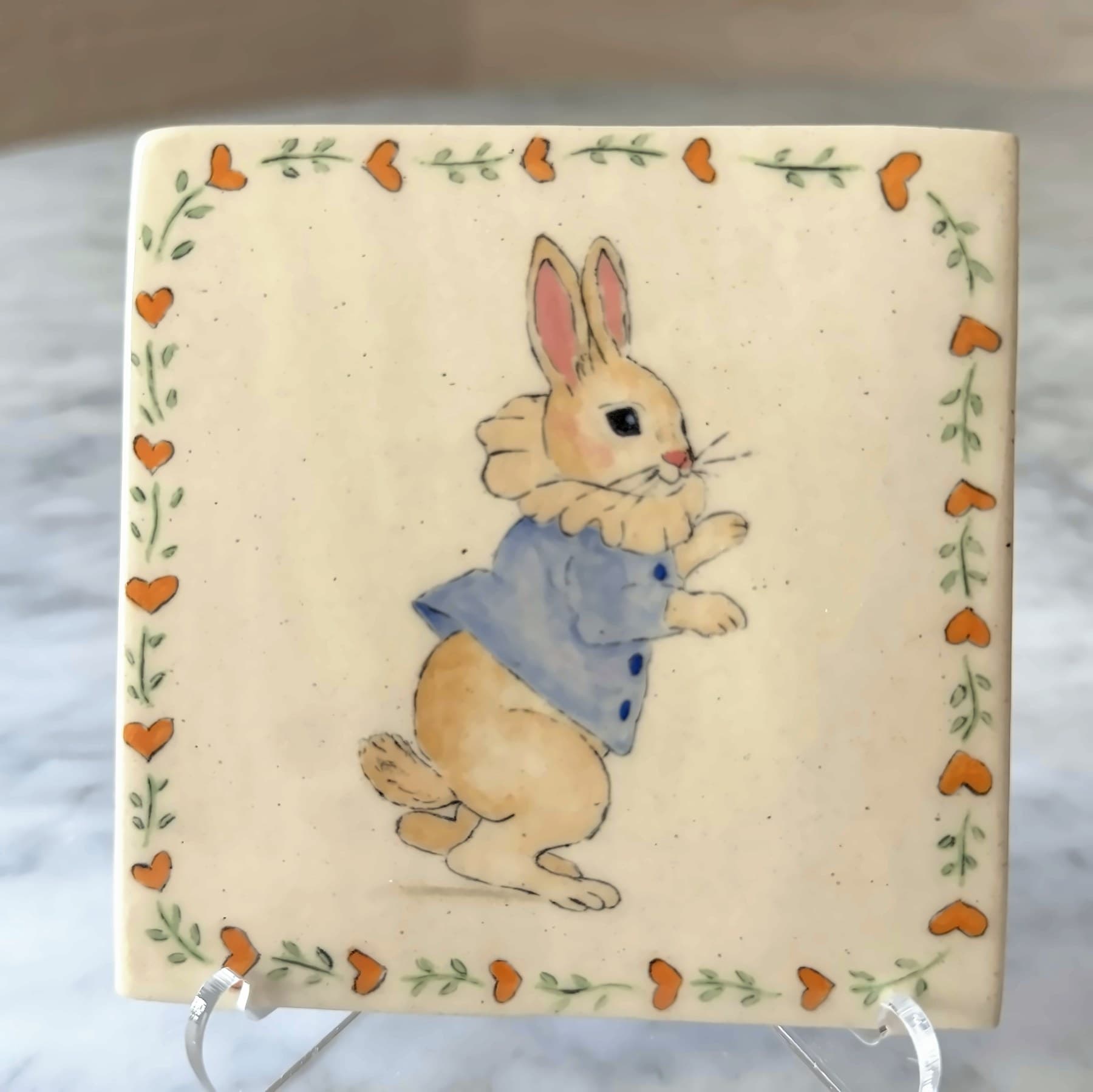 A tile painted with a rabbit in a blue coat and ruffled collar, framed by hearts and vines.
