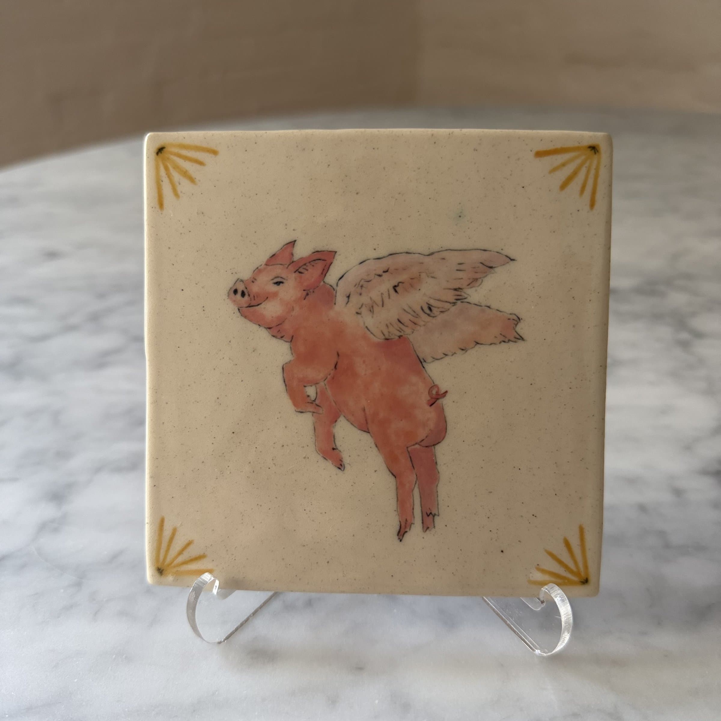 A hand-painted tile of a winged pig in mid-flight.