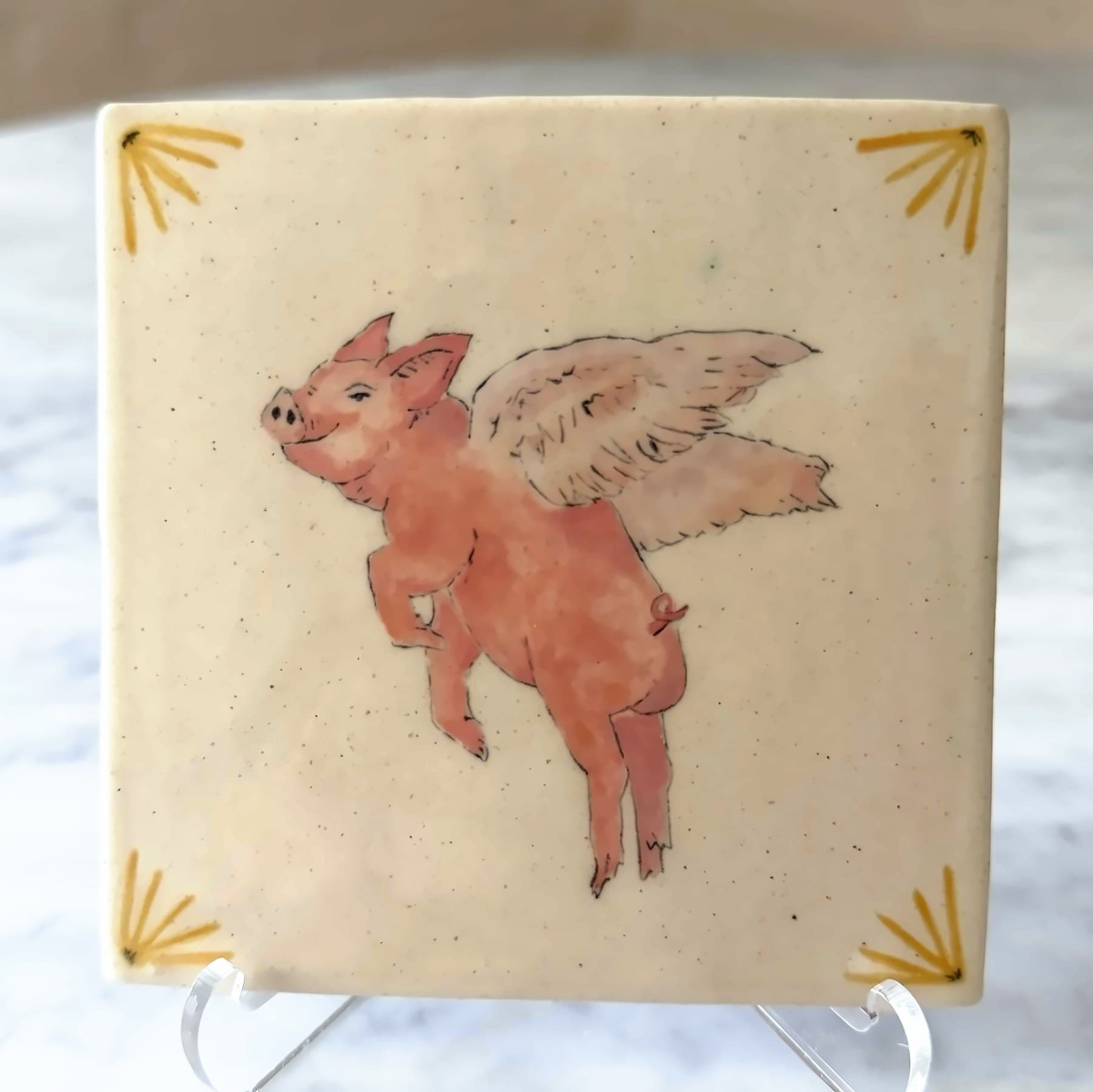 A tile painted with a smiling winged pig, sunbursts in the corners.