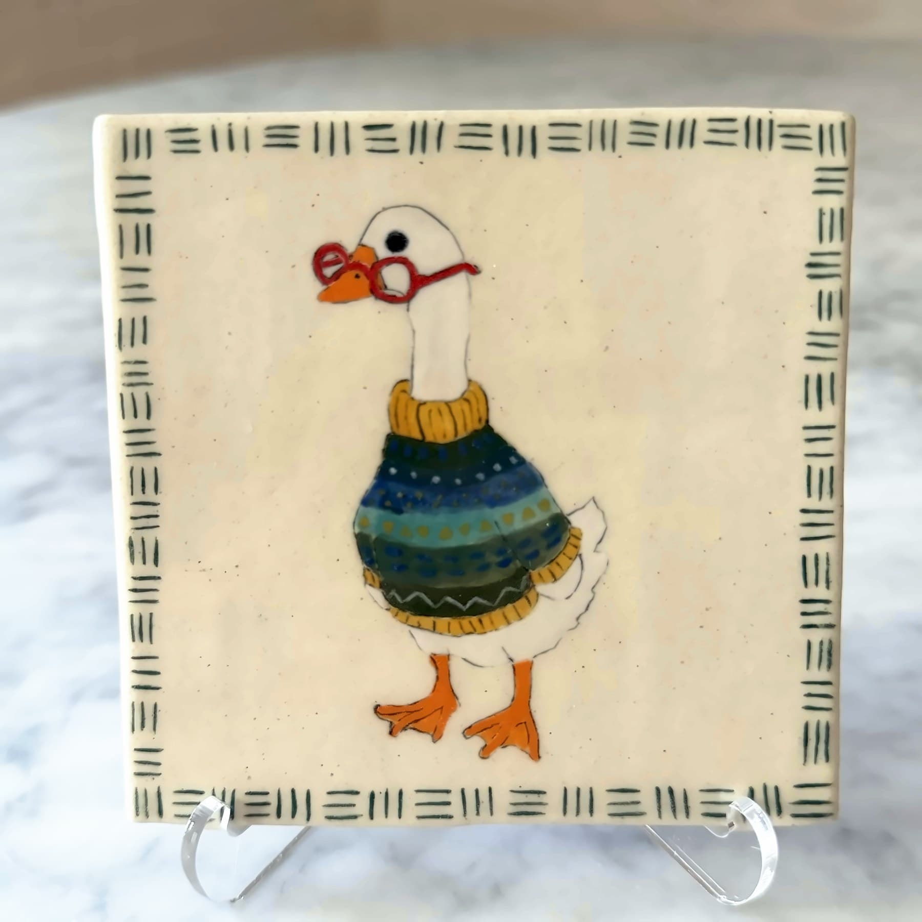 A tile painted with a white goose in red glasses and a striped sweater.