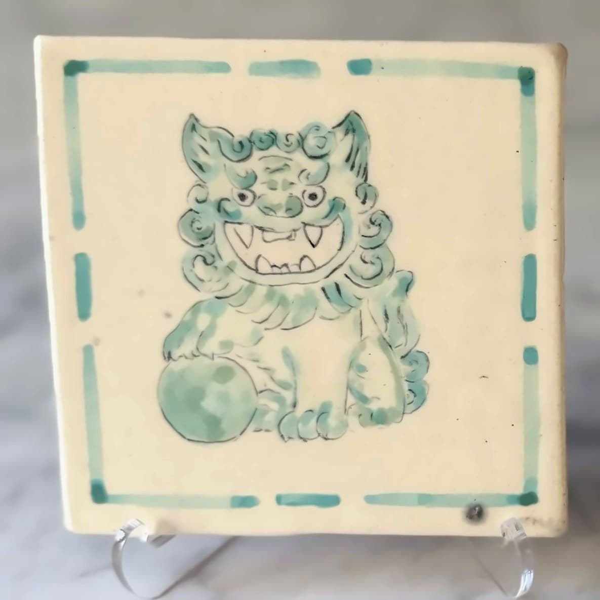 A tile painted in green with a roaring foo dog resting a paw on a ball.