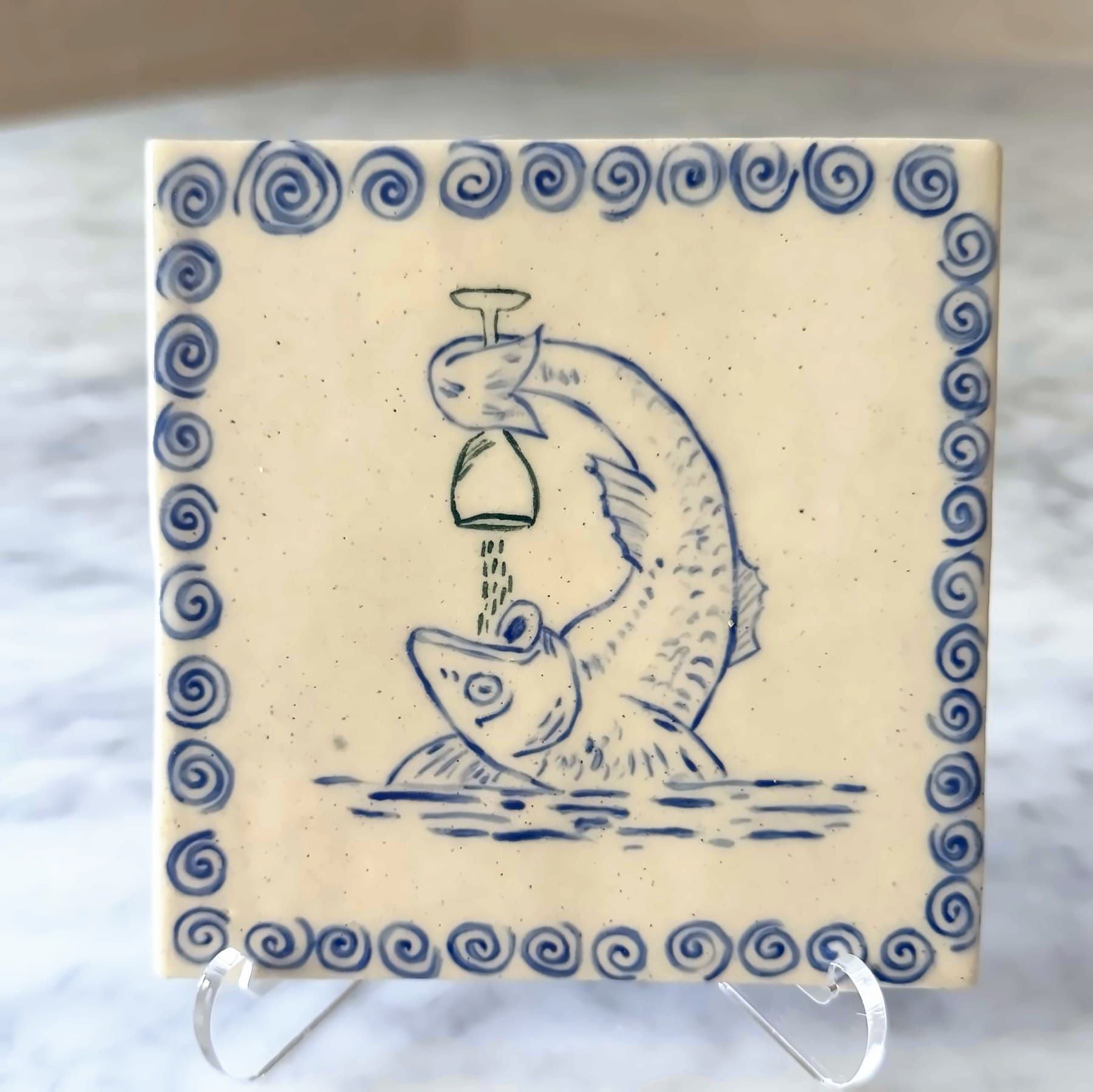 A blue tile drawing of a leaping fish tipping a wine glass into its mouth, framed by swirls.