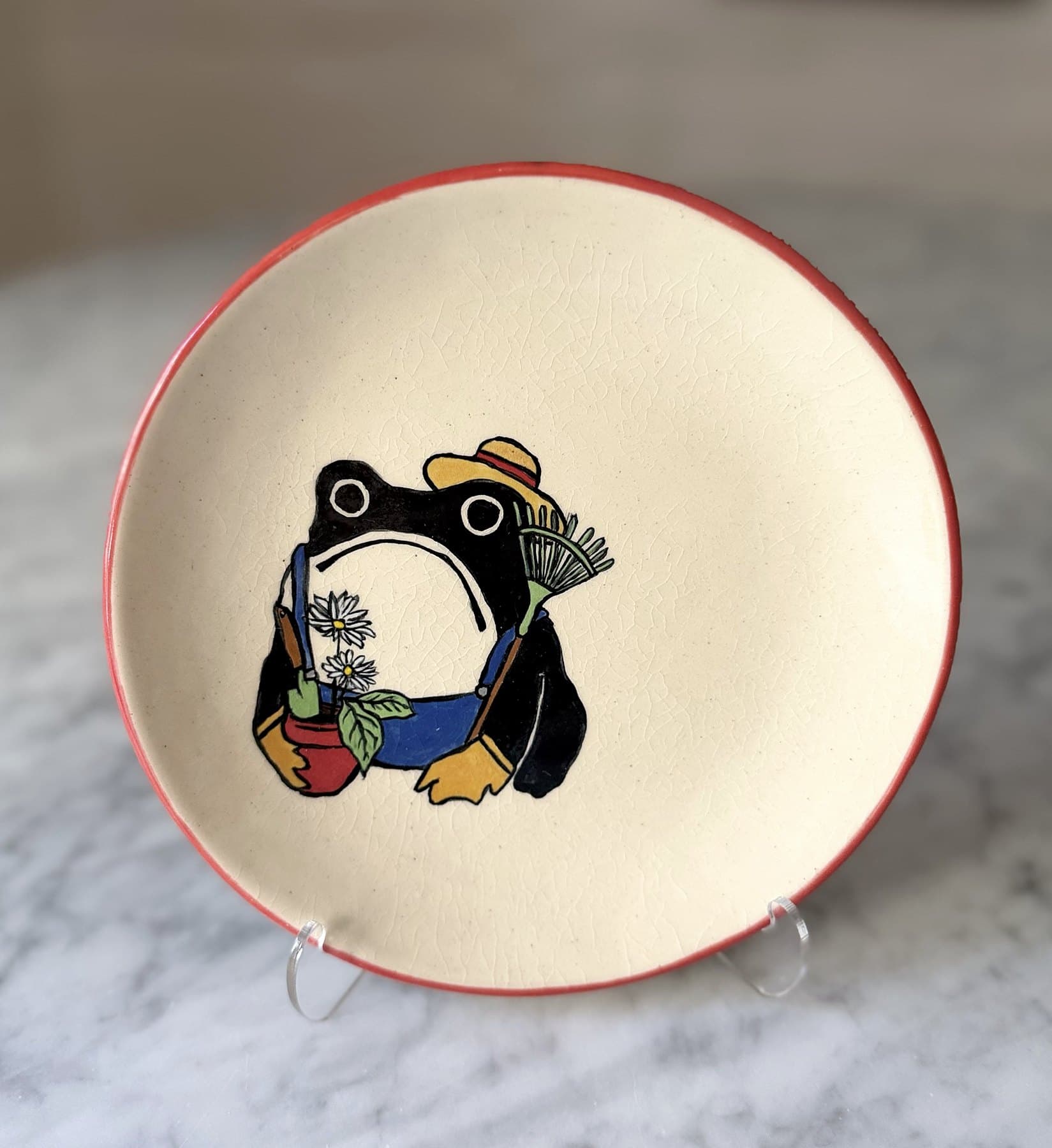 A red-rimmed plate painted with a frowning frog in overalls and a straw hat.