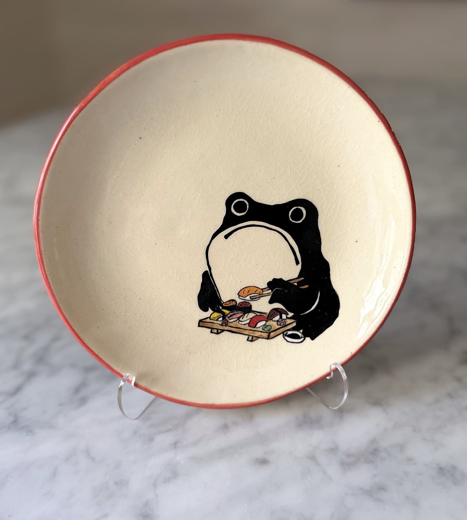 A red-rimmed plate painted with a frowning black frog eating sushi from a wooden board.
