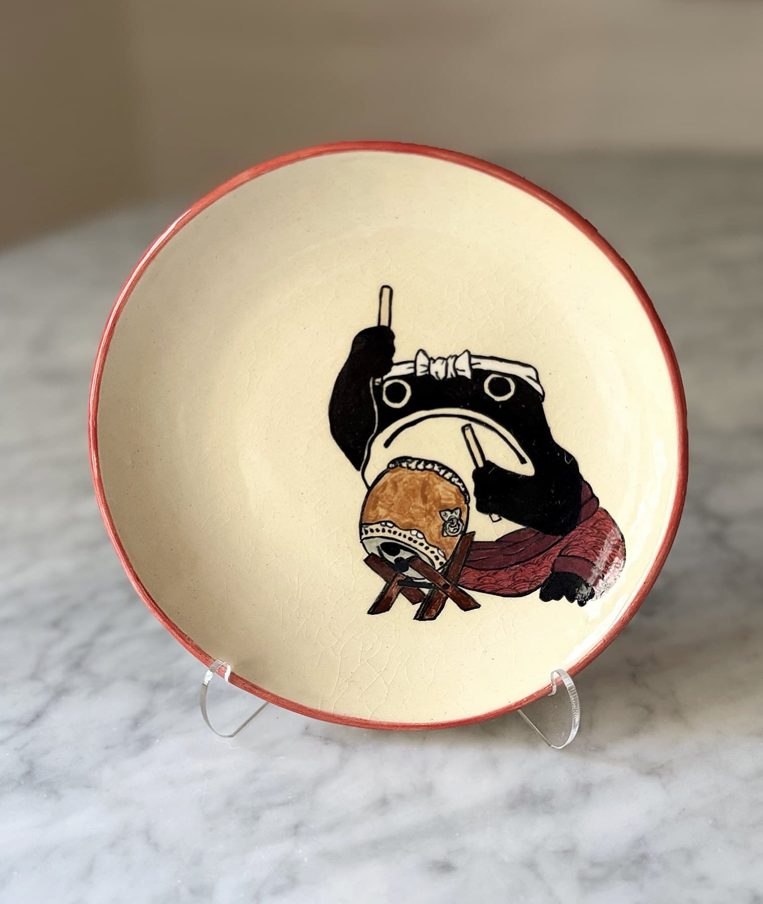 A red-rimmed plate painted with a frog in a white headband drumming a taiko drum.