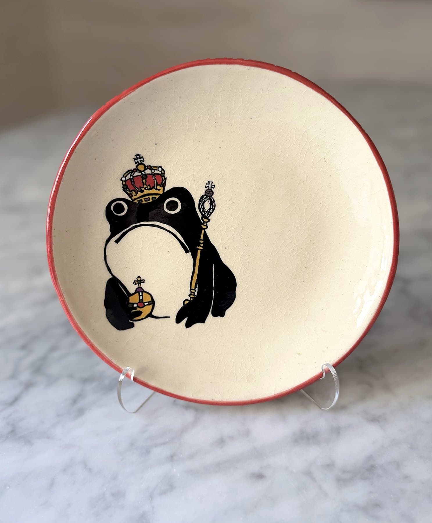 A red-rimmed plate painted with a frowning frog wearing a jeweled crown, holding an orb and sceptre.