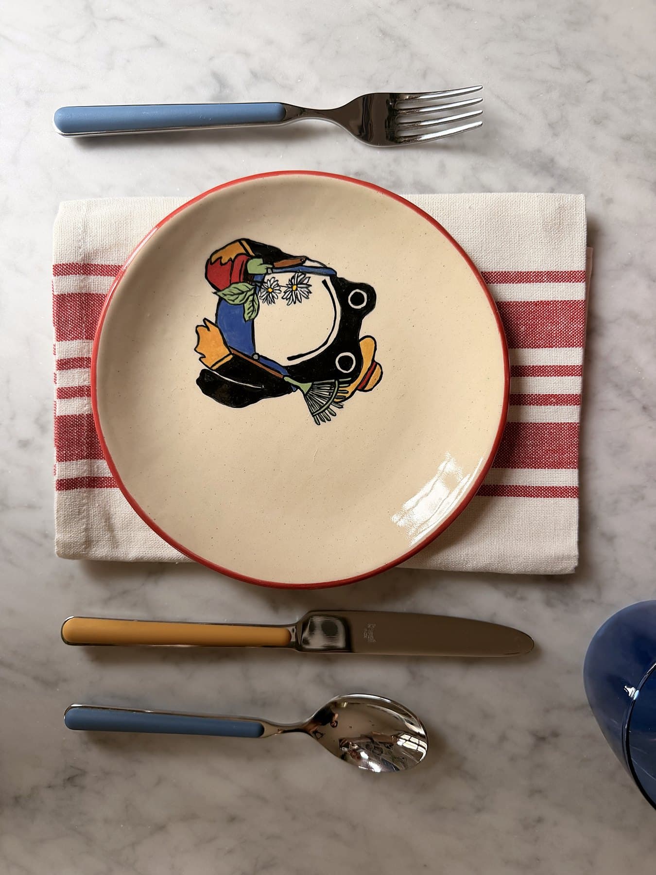 A red-rimmed frog plate set on a striped linen napkin with blue and yellow handled cutlery, seen from above.